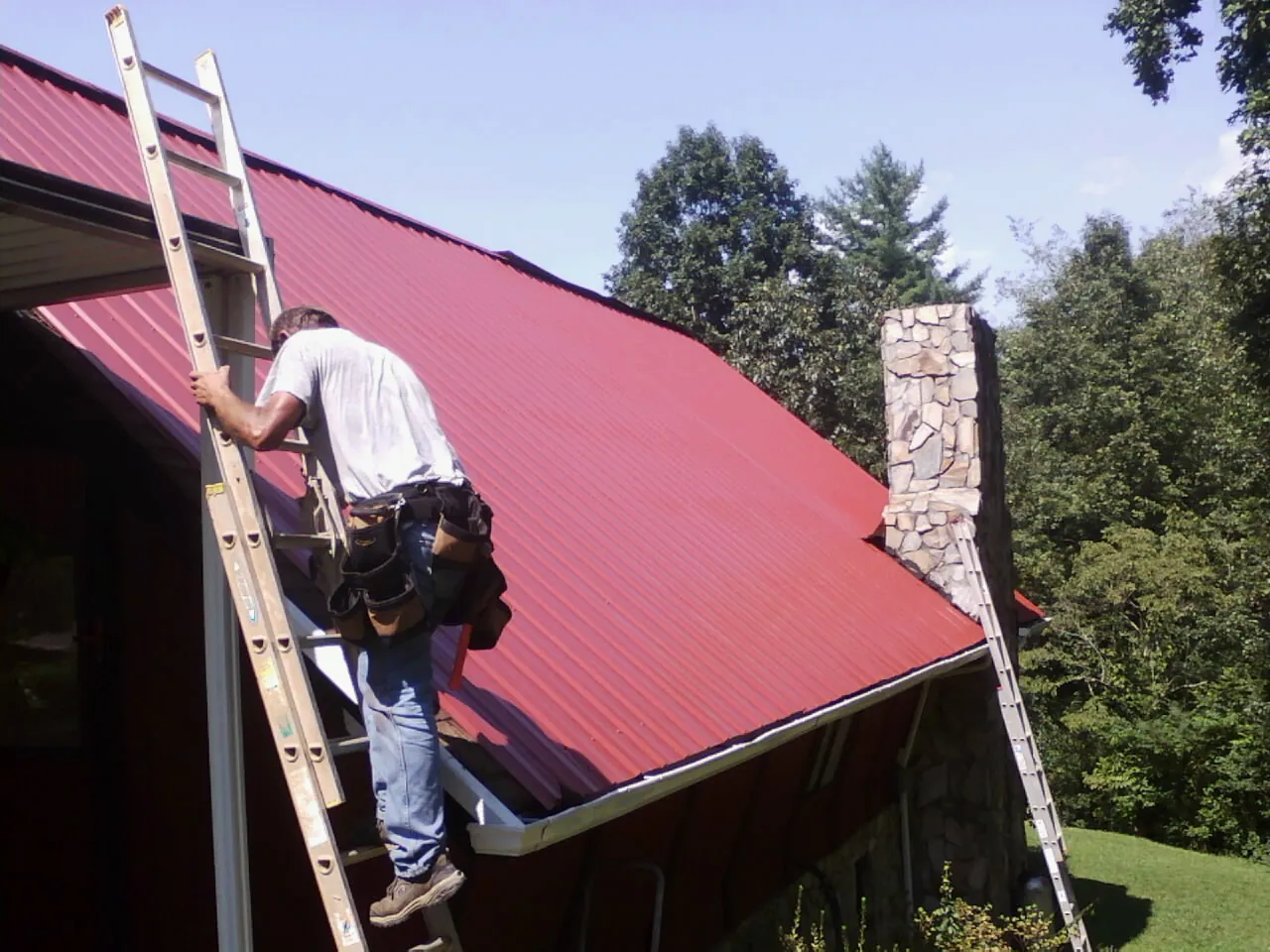 Roofer on a ladder beside a red metal roof and stone chimney — Soffit & Fascia Repair by Roofing Brothers in Pittsburg